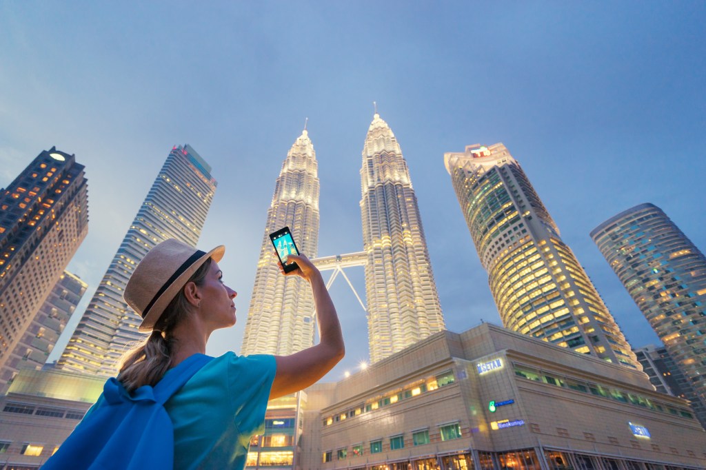 Travel and technology. Young  woman taking photo with her smartphone of Petronas Twins Towers in Kuala-Lumpur at evening, Malaysia, 23 November 2015.