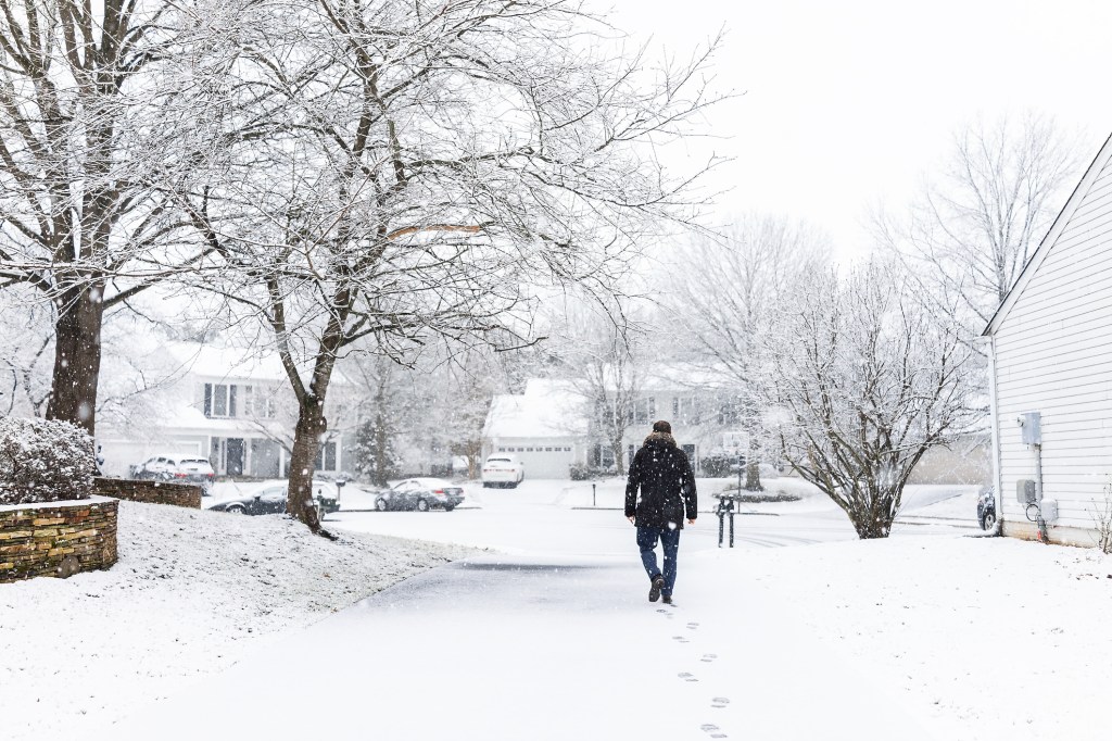 Man walking in driveway in neighborhood with snow covered ground during blizzard white storm, snowflakes falling in Virginia suburbs, single family homes to check mail in mailbox