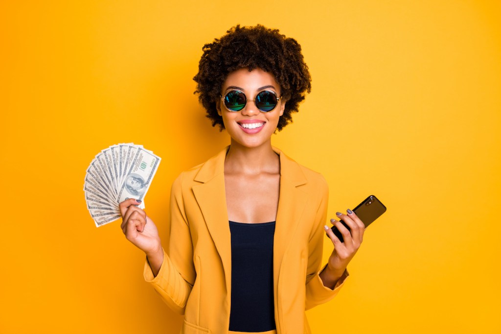 Portrait of positive cheerful afro american lady freelancer get cashback salary hold money fan use smartphone buy purchase pay online wear style blazer suit isolated over yellow color background